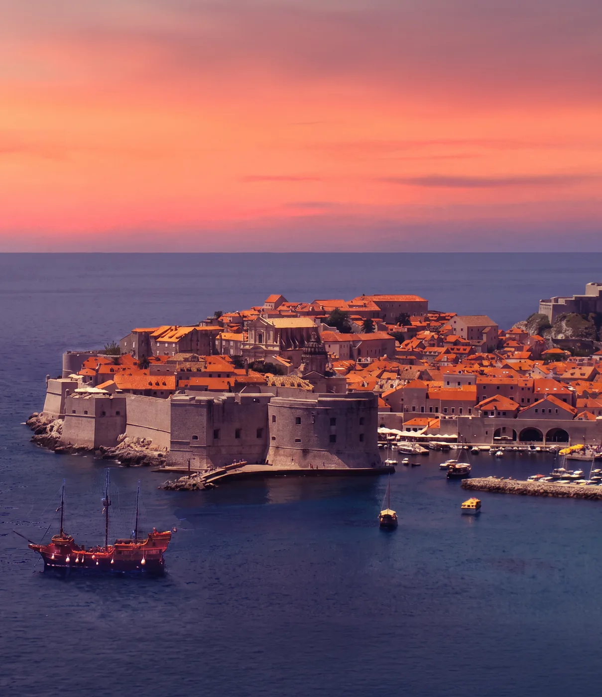 The walled old town of Dubrovnik at dusk under a rose sky, with a sailing ship below